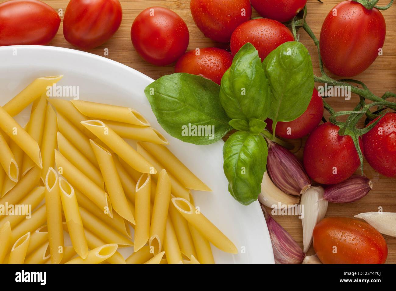 assiette de pâtes de blé dur, format penne lisse, sur une planche à découper en bois avec tomates mûres, feuilles de basilic frais et gousses d'ail Banque D'Images