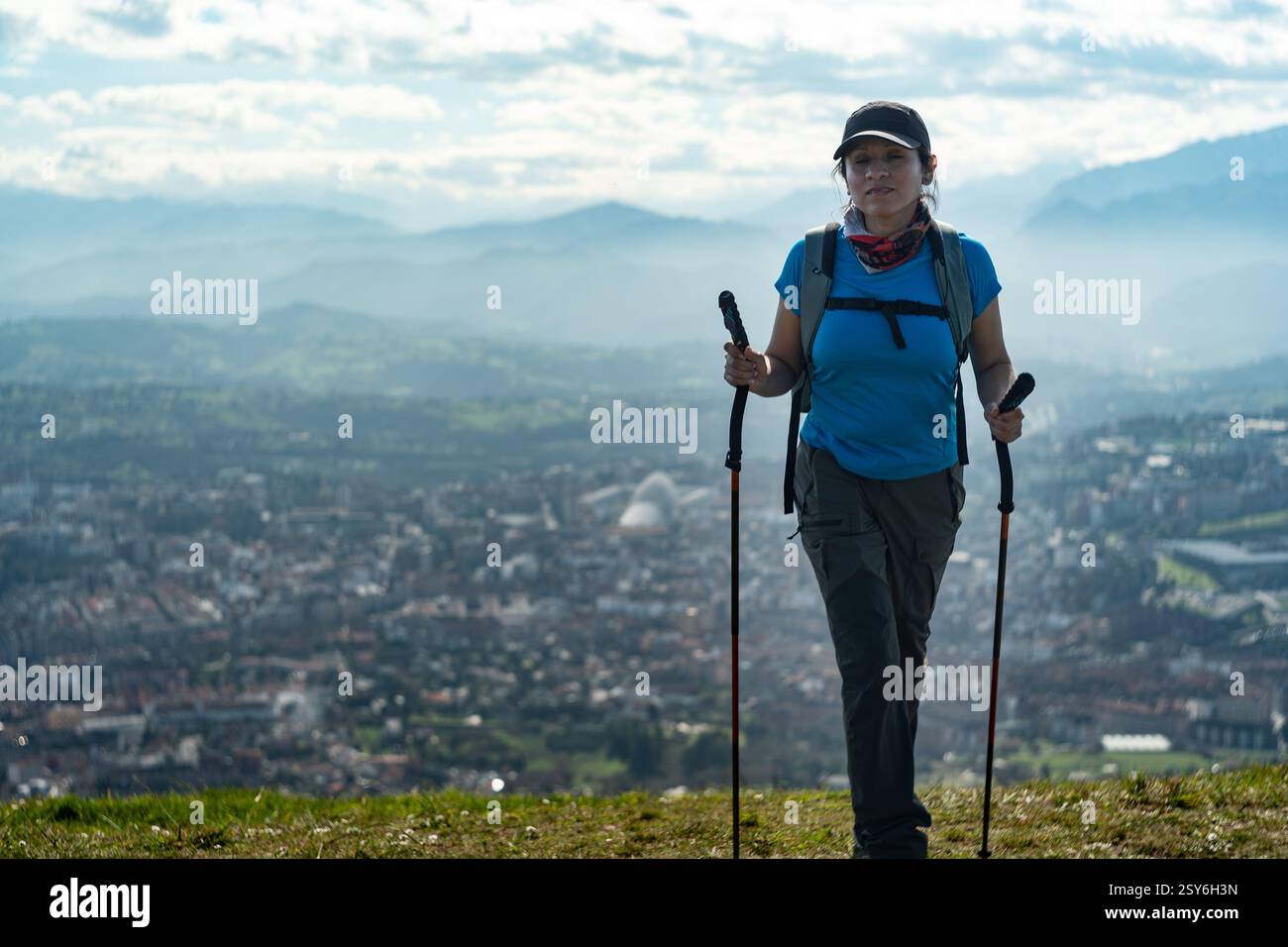 Femme atteint le sommet de la montagne Naranco tenant deux bâtons de randonnée et un fond impressionnant derrière le ciel, les montagnes et la ville. Banque D'Images