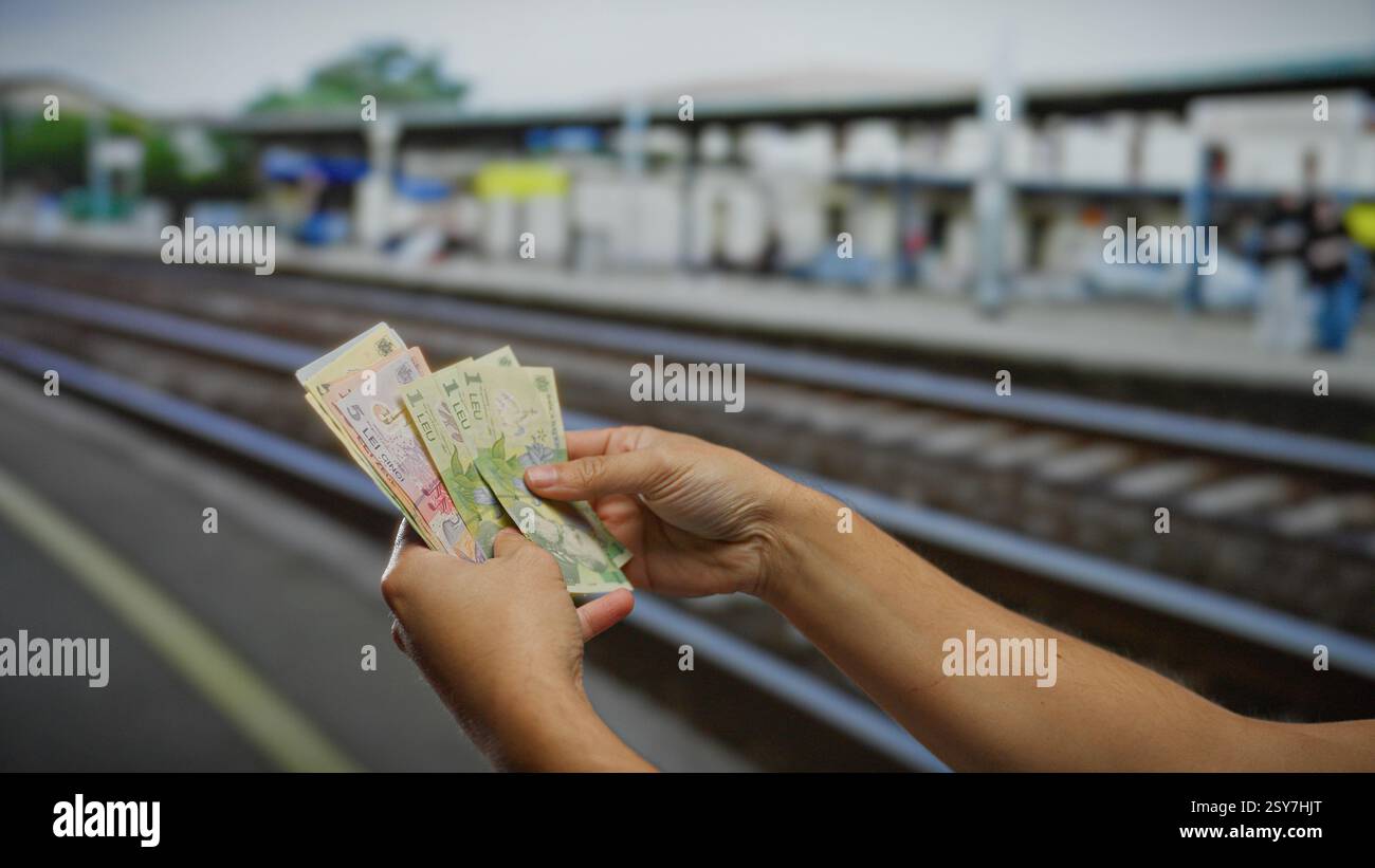Homme tenant des billets de banque en leu roumains dans une gare, mettant en évidence les thèmes du voyage et de la finance dans un cadre extérieur. Banque D'Images