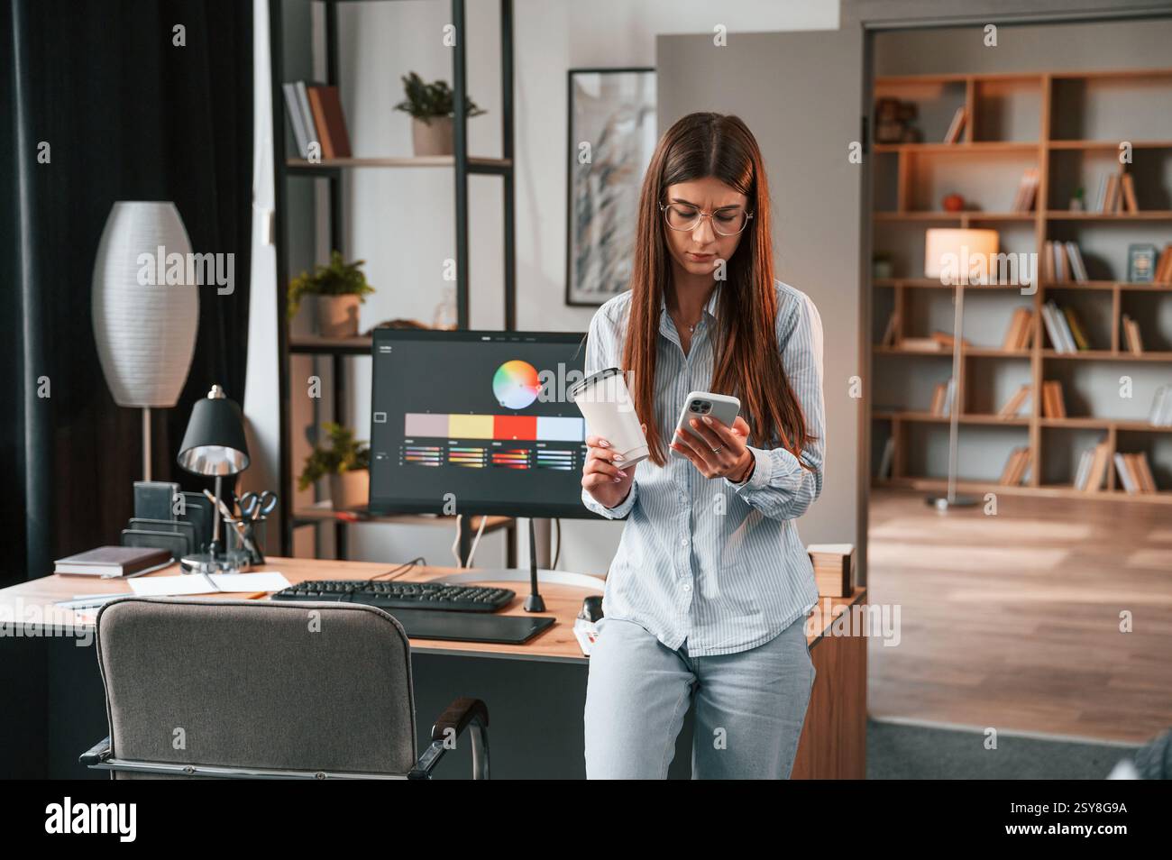 Faire une pause, se tenir debout avec un smartphone et une tasse de boisson dans les mains. Jeune femme designer est dans le bureau moderne. Banque D'Images