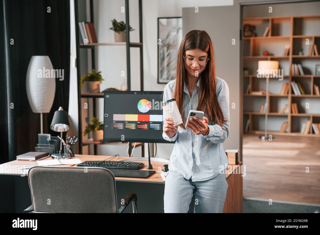 Faire une pause, se tenir debout avec un smartphone et une tasse de boisson dans les mains. Jeune femme designer est dans le bureau moderne. Banque D'Images