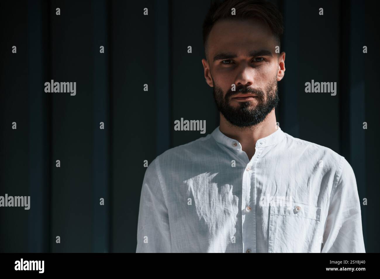 Jeune homme beau avec la barbe et en chemise blanche se tient debout sur fond vert foncé. Banque D'Images