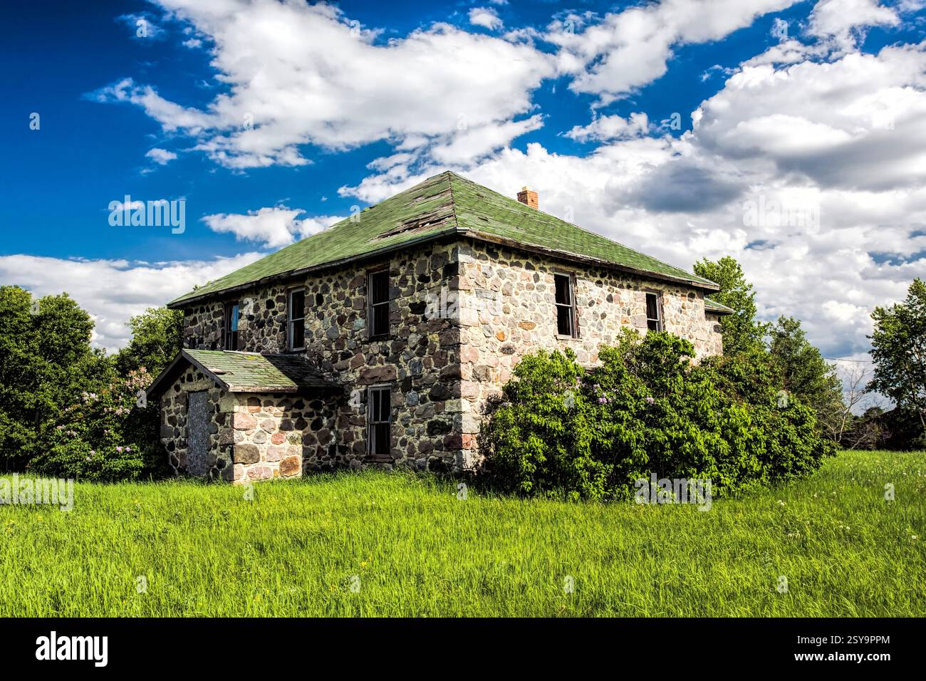 Grande maison en pierre avec un toit vert. Il manque des tuiles au toit. La maison est entourée d'un grand champ herbeux Banque D'Images