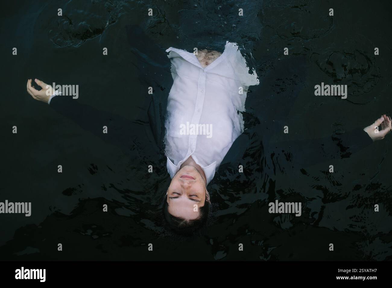 Un homme vêtu d'un costume flotte sur l'eau sombre avec ses bras tendus, les yeux fermés, et une expression paisible. La scène surréaliste transmet un sens de Banque D'Images
