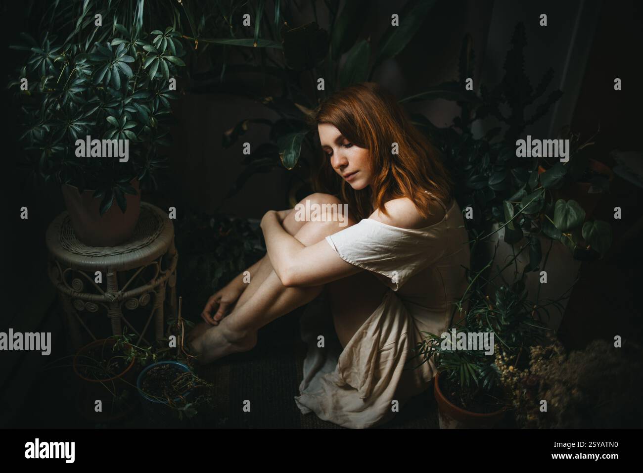 Une jeune femme s'assoit paisiblement parmi les plantes d'intérieur luxuriantes, entourée de verdure. Son expression sereine et sa posture détendue évoquent un sentiment de tranquillité Banque D'Images