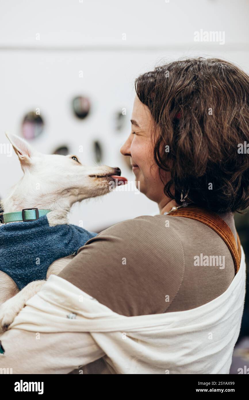 Une jeune femme partage un moment joyeux avec son chien qui lèche son visage de manière ludique dans une librairie. Capturée dans un cadre chaleureux et intérieur, cette image transmet Banque D'Images