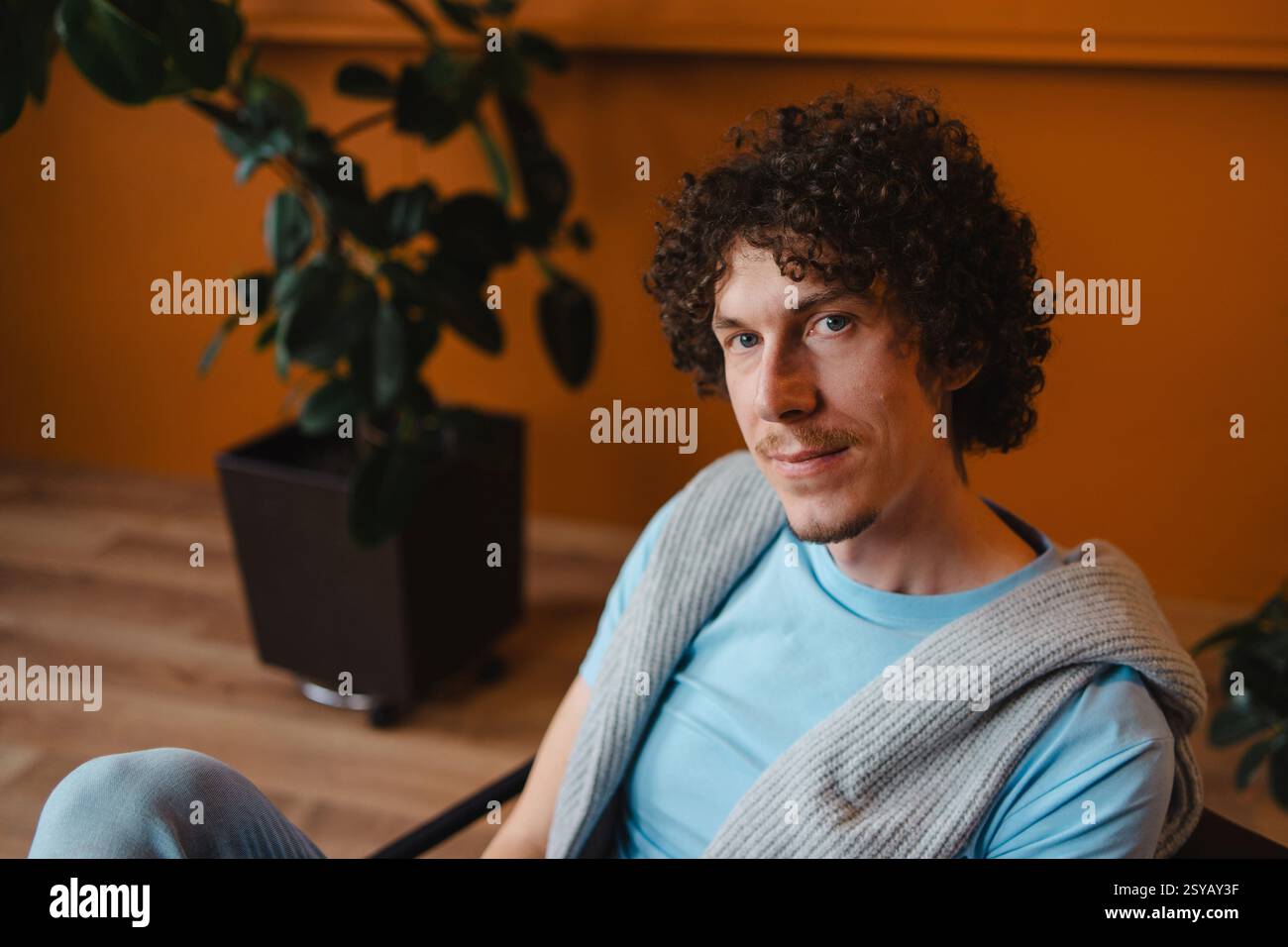 Jeune homme aux cheveux bouclés dans un cadre confortable, portant une chemise bleue et un pull en tricot. Chambre aux tons chauds avec des plantes, créant un atmo détendu et naturel Banque D'Images
