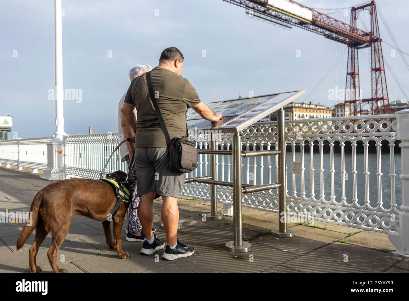 Un aveugle avec un chien-guide a lu un panneau d'information sur un pont surplombant une rivière pittoresque et des bâtiments lointains. La conception industrielle du pont ajoute Banque D'Images