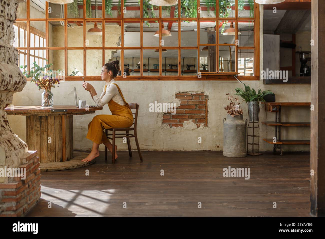 Une femme se livre au télétravail à l'aide d'un ordinateur portable sur une table circulaire en bois dans un café rustique et magnifiquement vieilli avec des murs de briques visibles et de grandes fenêtres Banque D'Images