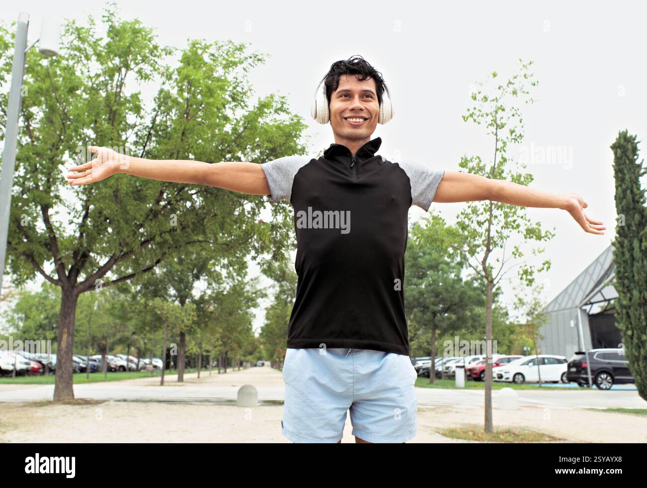 Athlète masculin latino joyeux dans un parc avec les bras écartés, portant des écouteurs, profitant du plein air et se sentant libre. Banque D'Images