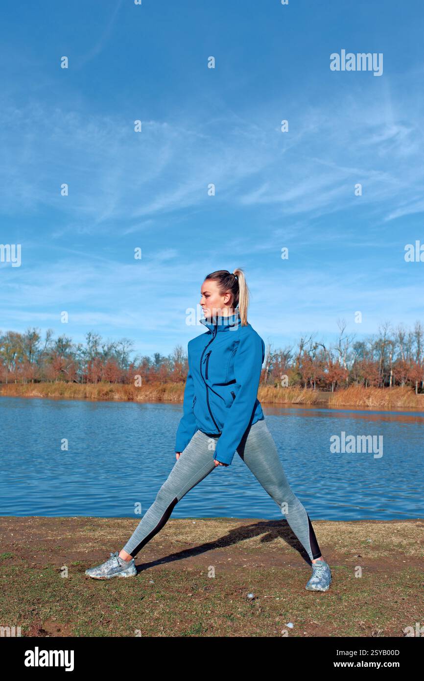 Femme en tenue athlétique s'étend près d'un lac tranquille sous un ciel bleu clair. Elle porte une veste bleue et des leggings, s'engageant dans l'exercice en plein air sur un Banque D'Images
