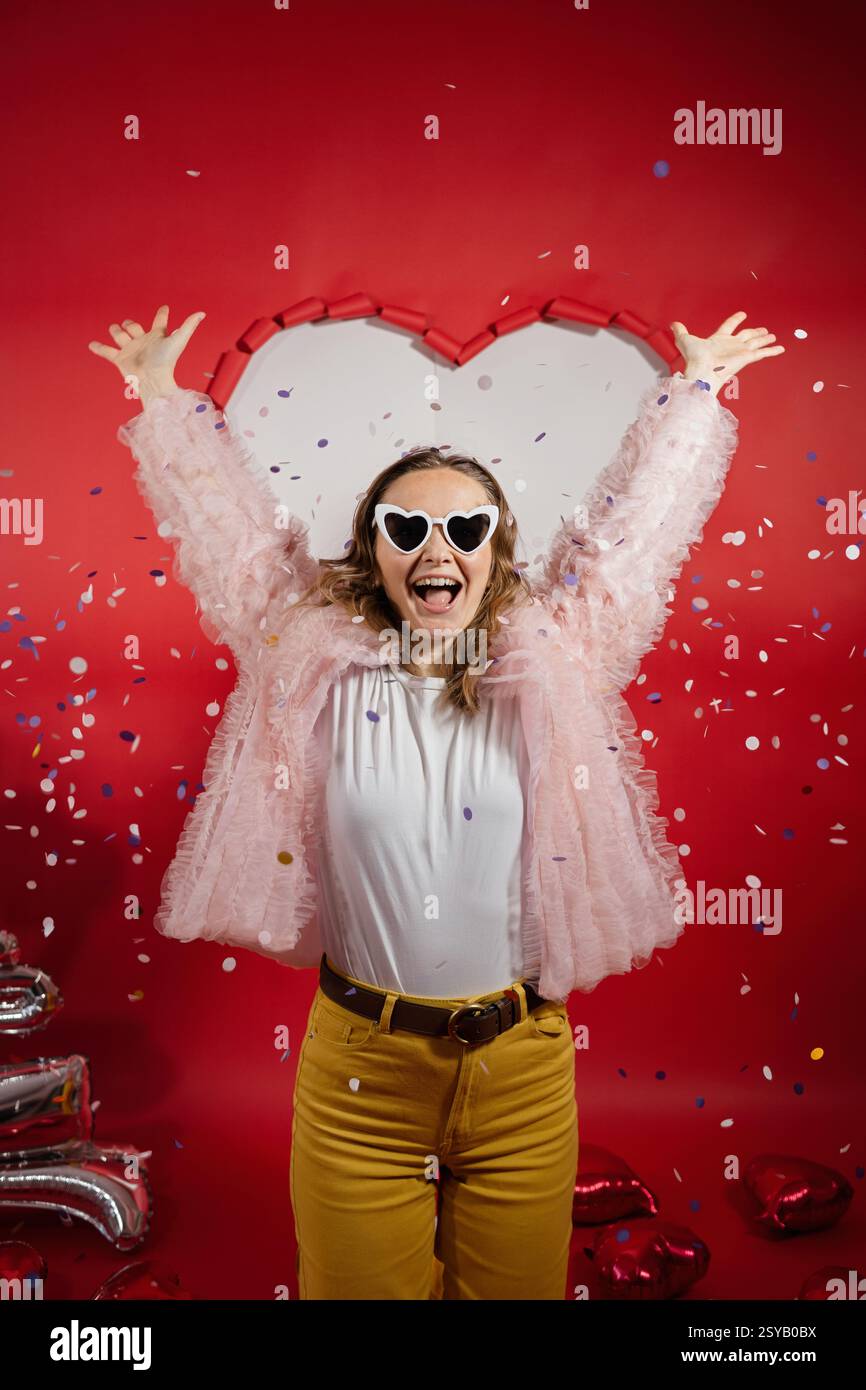 Une femme joyeuse jette ses bras en l'air au milieu de confettis tombant, célébrant la Saint-Valentin. Elle porte des lunettes de soleil en forme de cœur et une tenue festive Banque D'Images
