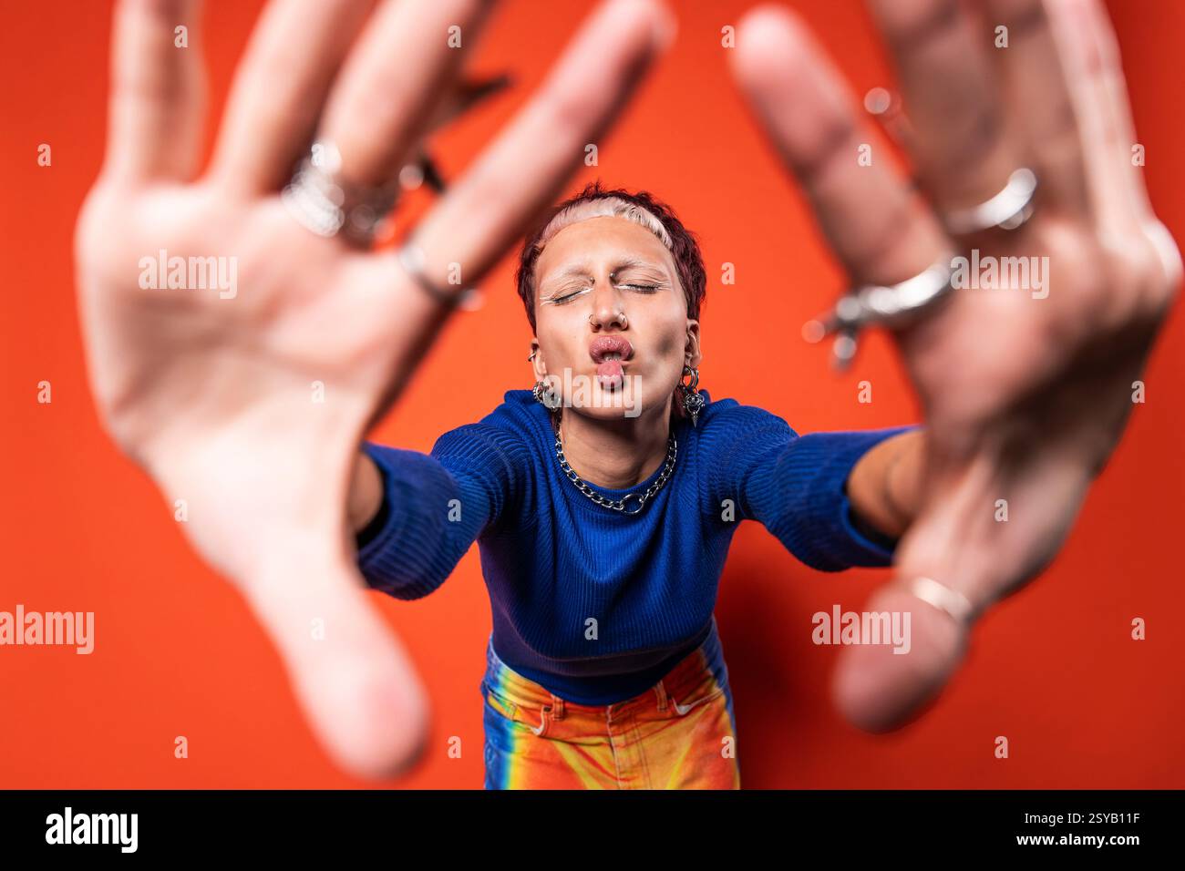 Une femme à la mode se démarque dans un pantalon vibrant, faisant un visage de poisson ludique. Elle présente des bijoux éclectiques et un haut bleu audacieux contre un brillant Banque D'Images