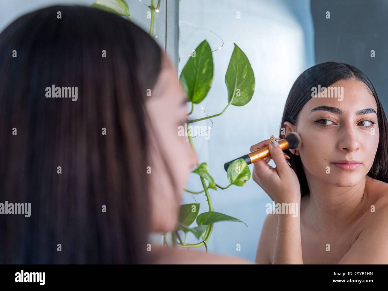 Une femme se maquille avec un pinceau tout en regardant dans un miroir. Les plantes vertes ajoutent une touche naturelle au cadre serein, créant une atmosphère apaisante Banque D'Images