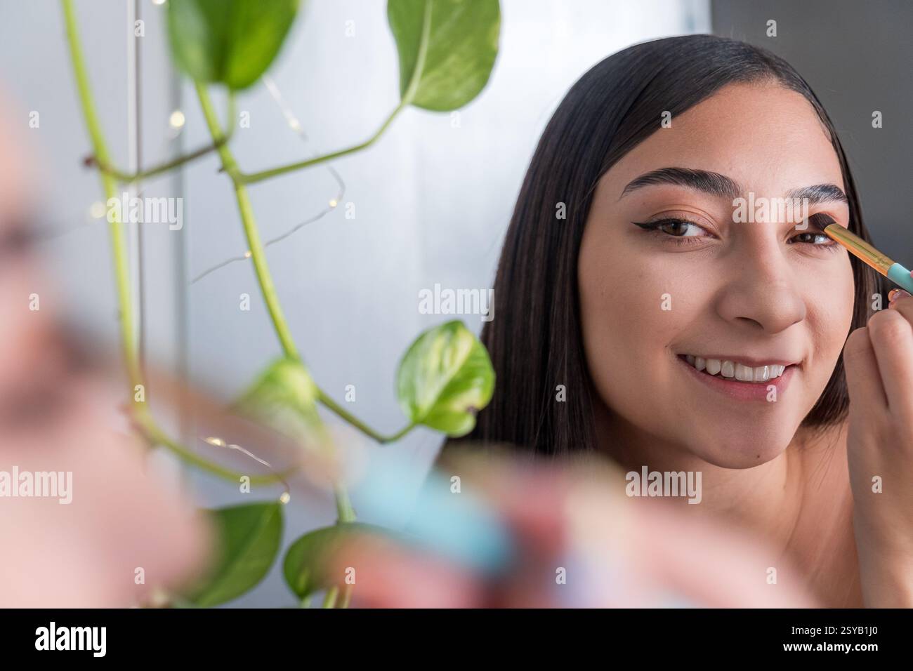 Une femme est vue souriante tout en appliquant le maquillage des yeux devant un miroir pour la séance de maquillage. Une plante verte est visible, ajoutant une touche de nature à t Banque D'Images
