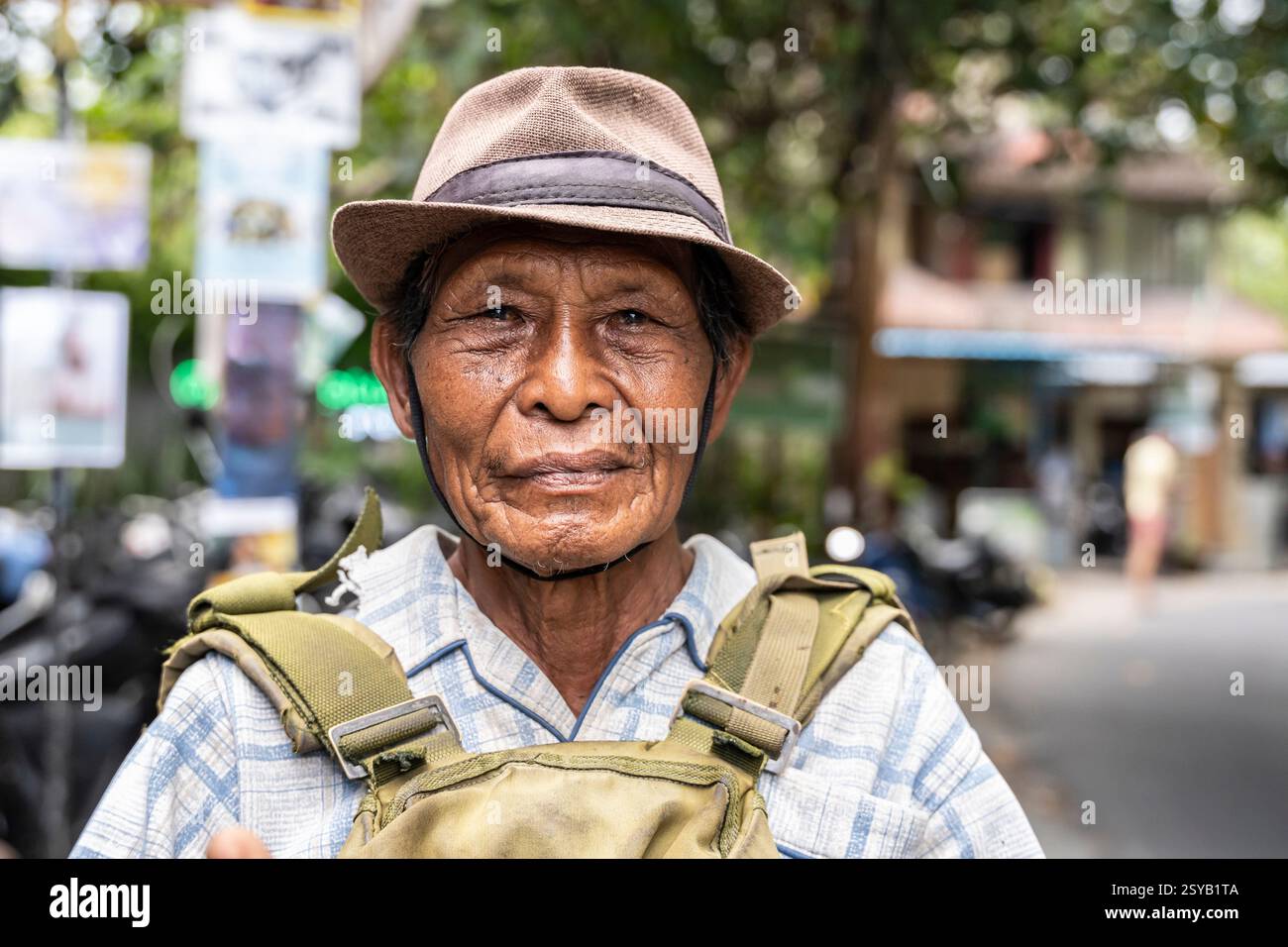 Un homme balinais âgé portant un chapeau et une chemise à carreaux se tient dehors, regardant dans la caméra avec une expression sereine. L'arrière-plan est flou Banque D'Images