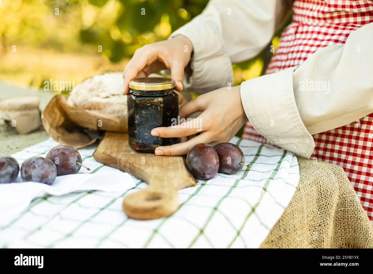Une personne prépare des canapés en utilisant des ingrédients frais. L'image comprend un pot de confiture, une miche de pain et des prunes disposées sur une planche à découper, posées sur un R. Banque D'Images