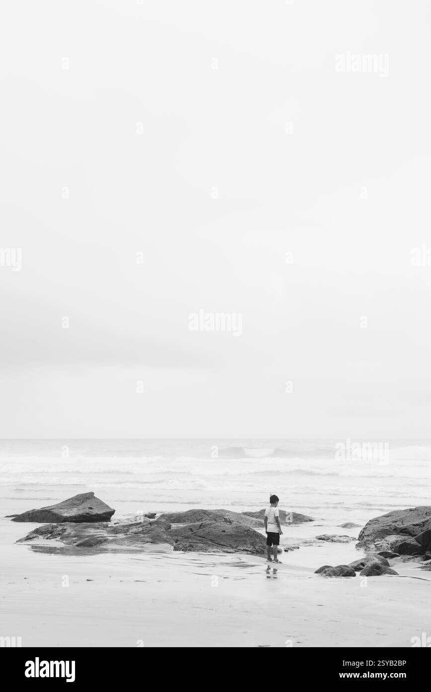 Un seul individu se tient sur une plage entourée de rochers, regardant l'océan. La photographie en noir et blanc capture un sentiment de solitude, de tranquillité Banque D'Images