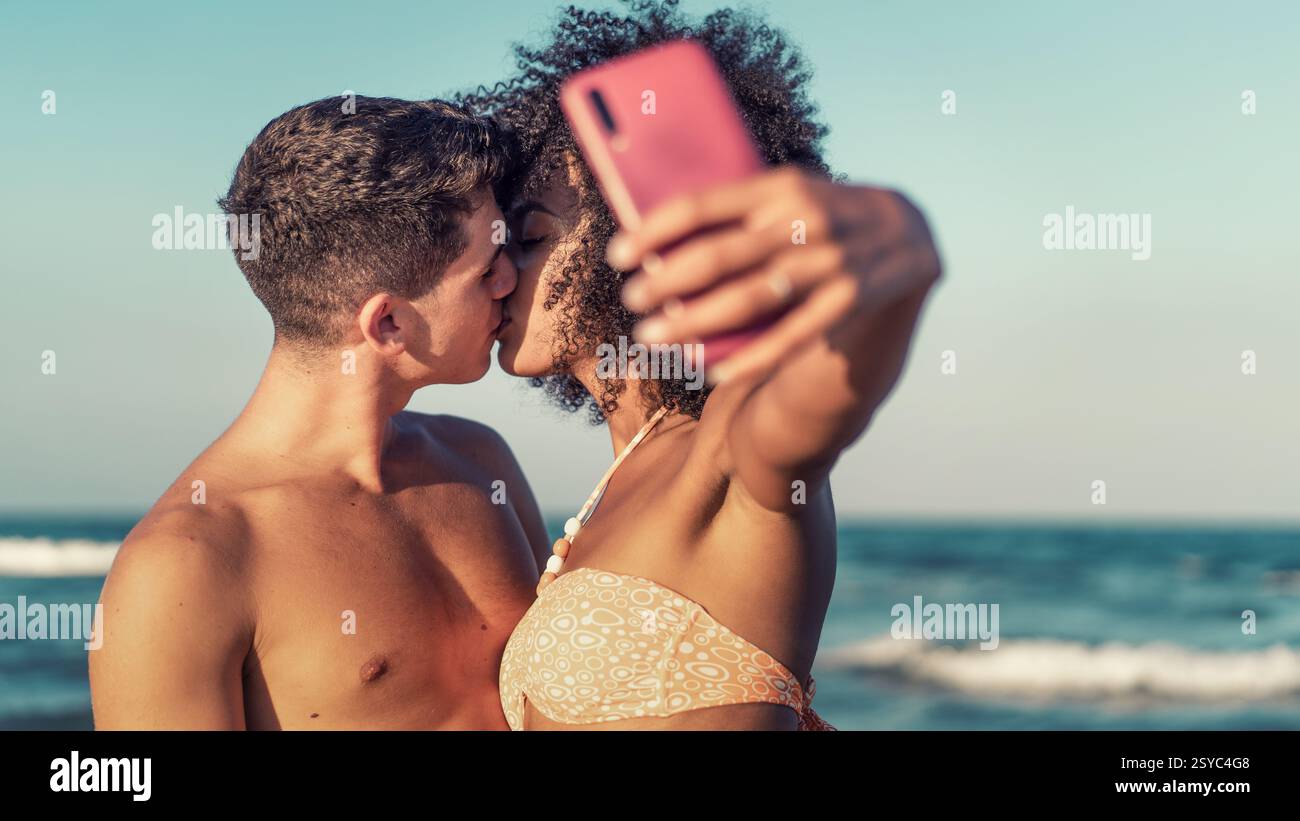 Jeune couple interracial s'embrassant tout en prenant un selfie sur la plage pendant l'été. Moment romantique et spontané capturant l'amour et le bonheur par le Banque D'Images