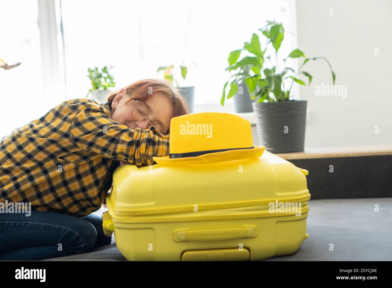 Voyageur épuisé reposant sur une valise emballée Une femme dans une chemise à carreaux jaune repose sa tête sur une valise jaune bien emballée, épuisée après Banque D'Images
