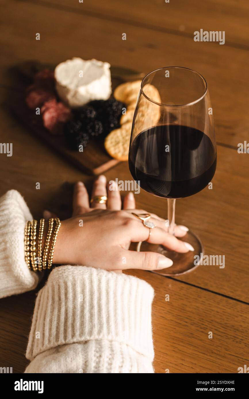 Une main dans un pull confortable et des bijoux en or repose près d'un verre de vin rouge et une planche de charcuterie sur une table en bois - élégante et accueillante. Banque D'Images