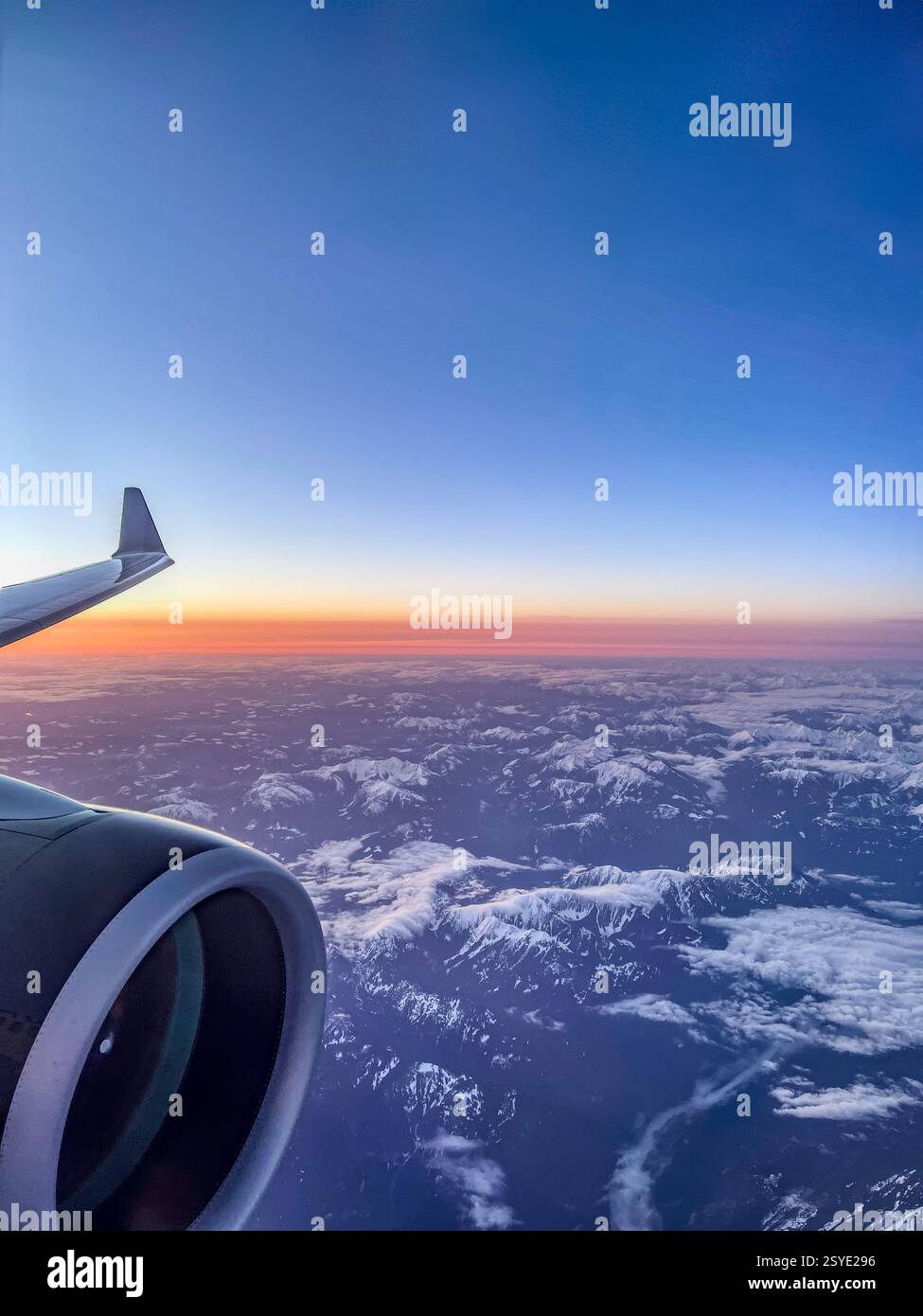 Vue aérienne depuis une fenêtre d'avion montrant l'aile et le moteur au-dessus d'une chaîne de montagnes enneigée au lever du soleil. Le ciel éclatant passe d'un bleu profond - Image de stock capturée avec un smartphone