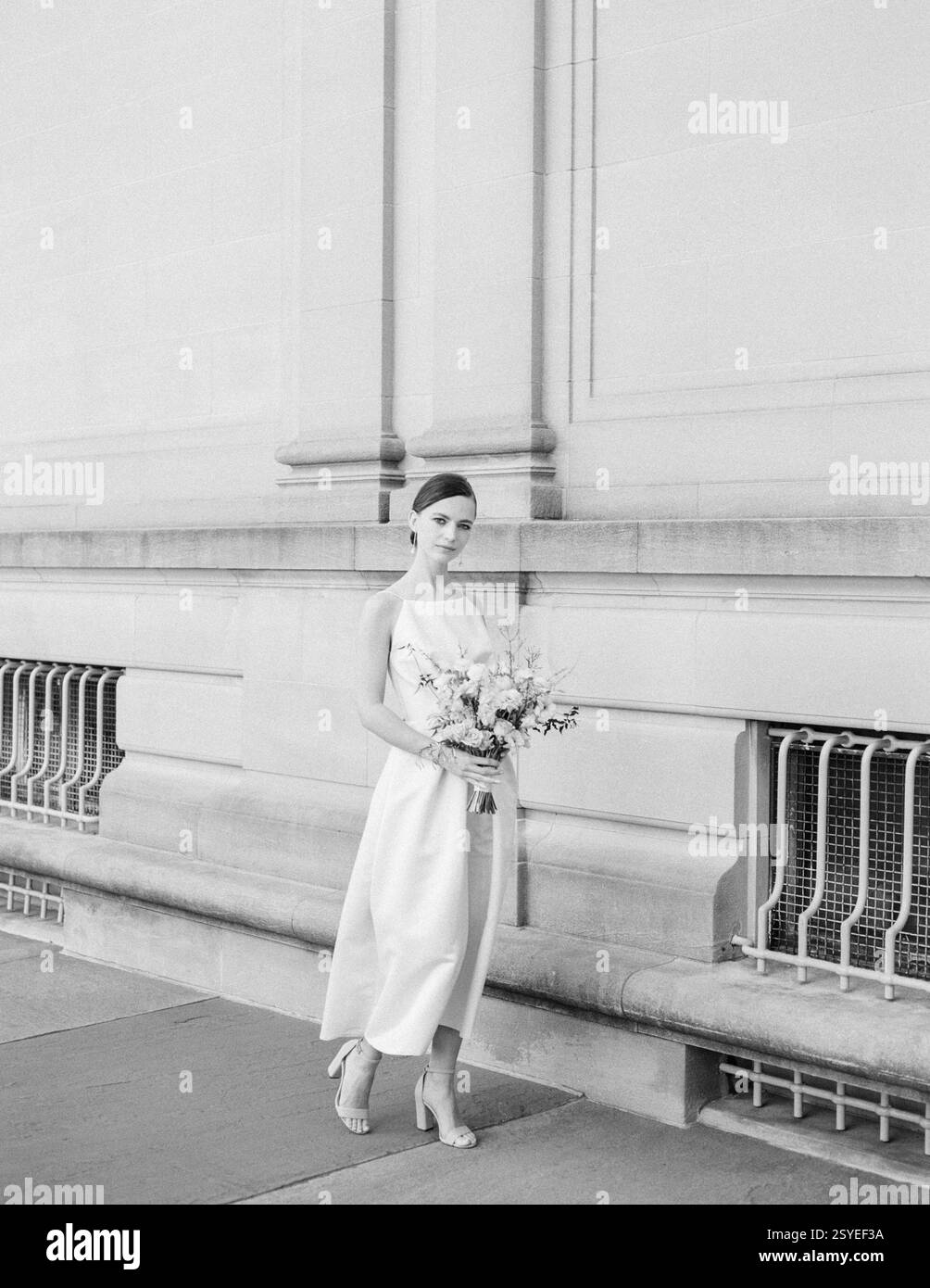 Mariée dans une robe blanche fluide pose gracieusement à l'extérieur d'une belle structure historique avec un bouquet de fleurs. Banque D'Images