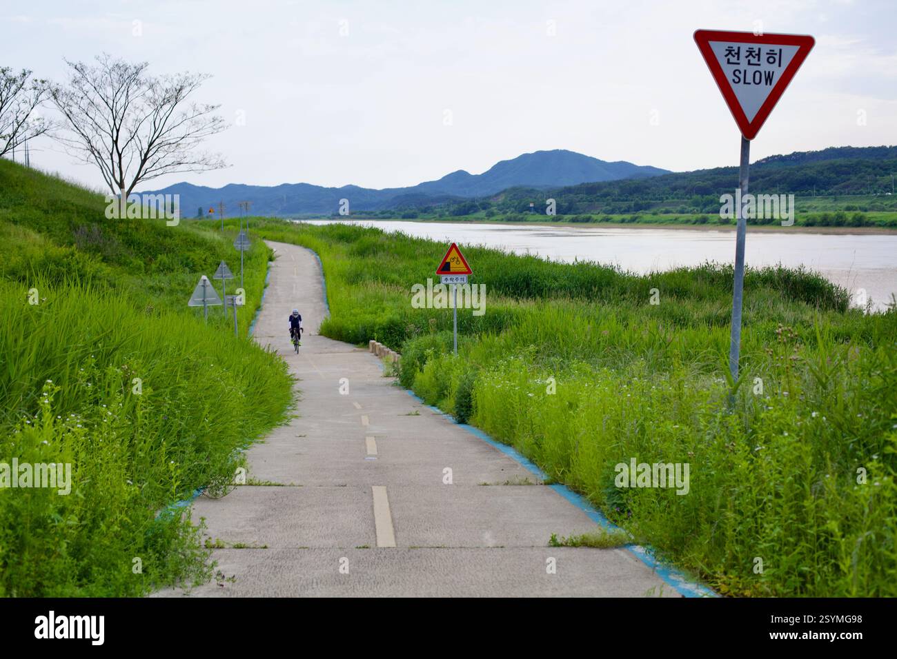 Gongju, Corée du Sud - 27 mai 2021 : un cycliste roule le long de la piste cyclable de la rivière Geum, passant un panneau «lent» avertissant d'une descente abrupte, surro Banque D'Images