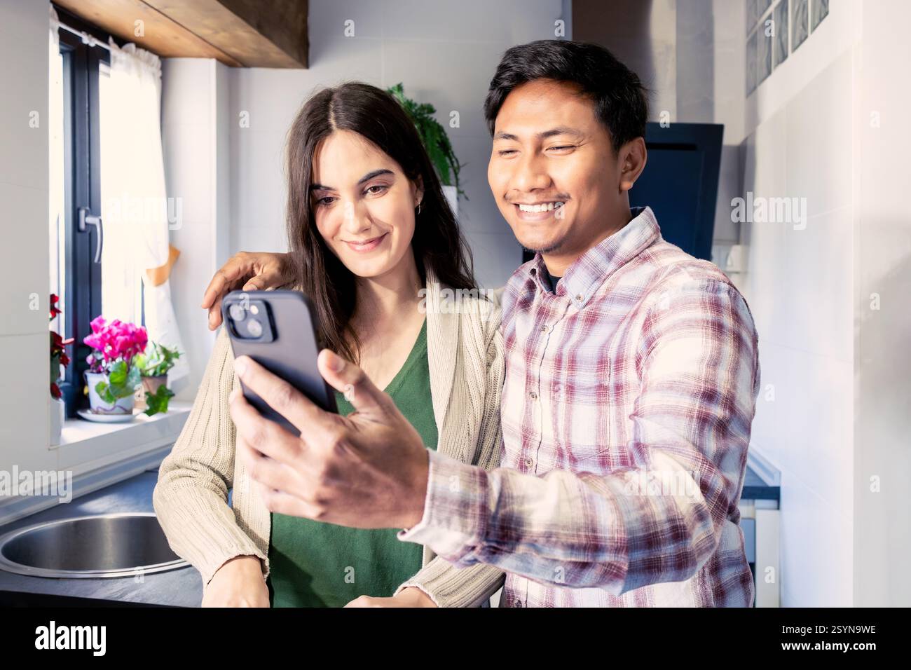 Couple souriant posant pour un selfie dans leur cuisine confortable, profitant d'un moment joyeux ensemble. la lumière naturelle, le décor chaleureux et les plantes en pot créent un r Banque D'Images