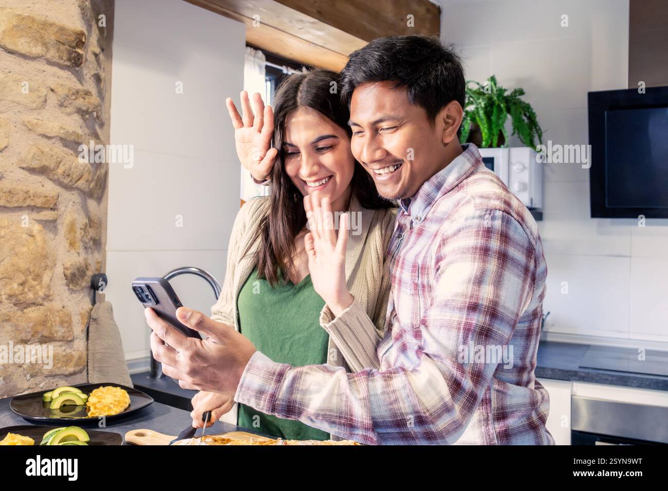 Couple joyeux souriant et agitant à leur smartphone tout en faisant un appel vidéo dans la cuisine. ils profitent d'un moment chaleureux et confortable ensemble pendant la préparation Banque D'Images