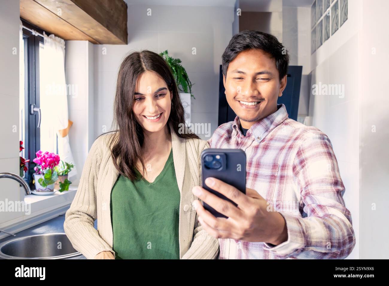 Couple souriant posant pour un selfie dans leur cuisine confortable, profitant d'un moment joyeux ensemble. la lumière naturelle, le décor chaleureux et les plantes en pot créent un r Banque D'Images