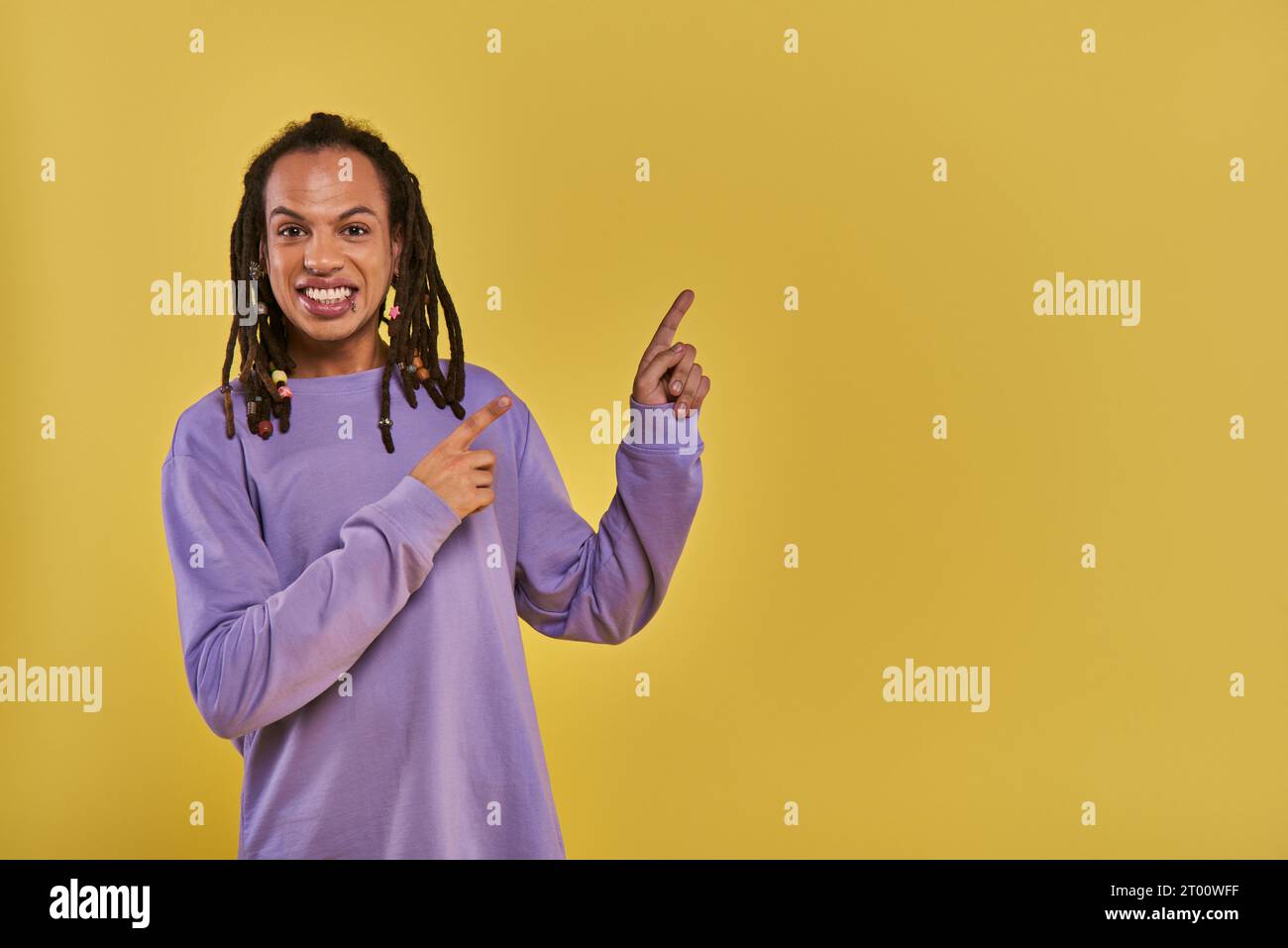 homme afro-américain joyeux avec dreadlocks percé lèvre pointant sur le côté droit sur fond jaune Banque D'Images