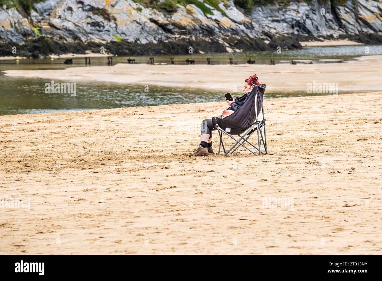 Une femme assise seule dans une chaise pliante et utilisant son téléphone portable sur Crantock Beach à Newquay en Cornouailles au Royaume-Uni. Banque D'Images