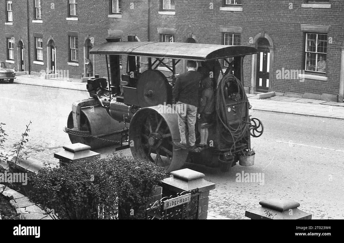 Années 1960, historique, un homme et son fils debout sur une plate-forme métallique de la cabine du conducteur d'un tracteur à vapeur à trois roues, ayant un tour sur elle pour voyager dans une rue de maisons mitoyennes victoriennes, Oldham Angleterre, Royaume-Uni. Banque D'Images