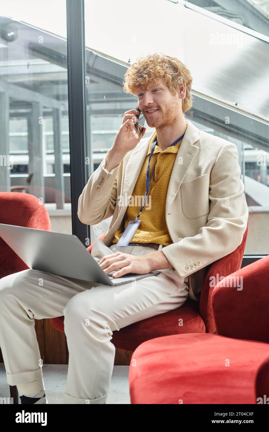 homme joyeux aux cheveux rouges tenant son ordinateur portable et parlant sur un téléphone portable avec toile de fond en verre, coworking Banque D'Images