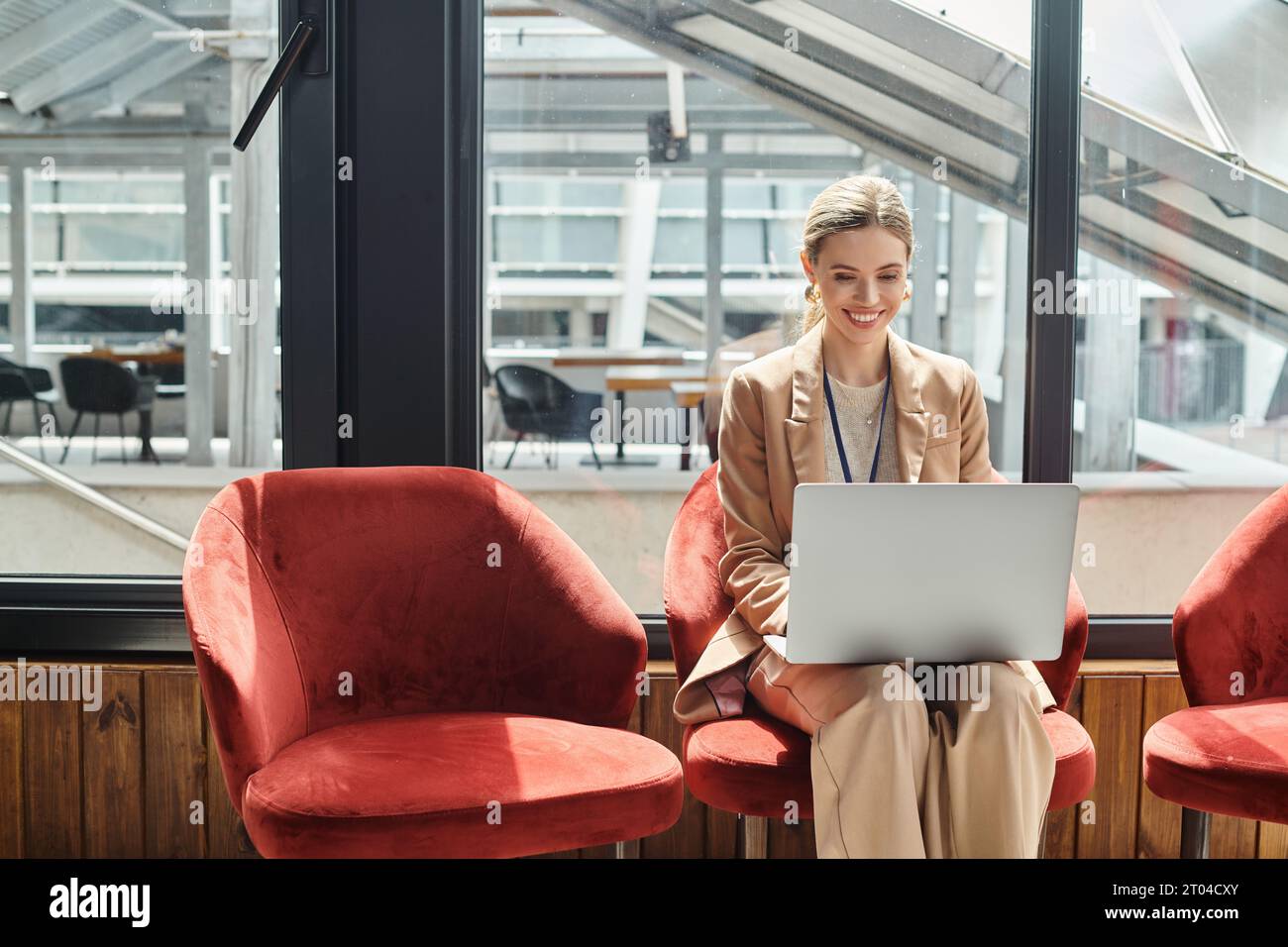 jeune employée blonde assise sur une chaise travaillant sur son ordinateur portable avec toile de fond en verre, concept de coworking Banque D'Images
