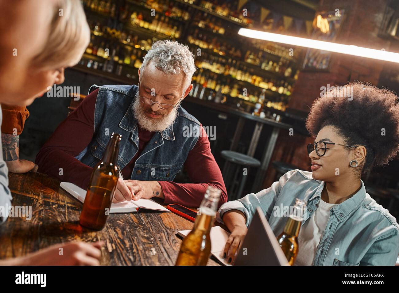 homme barbu d'âge moyen écrivant dans un cahier près de bouteilles de bière et collègues multiethniques dans un pub Banque D'Images