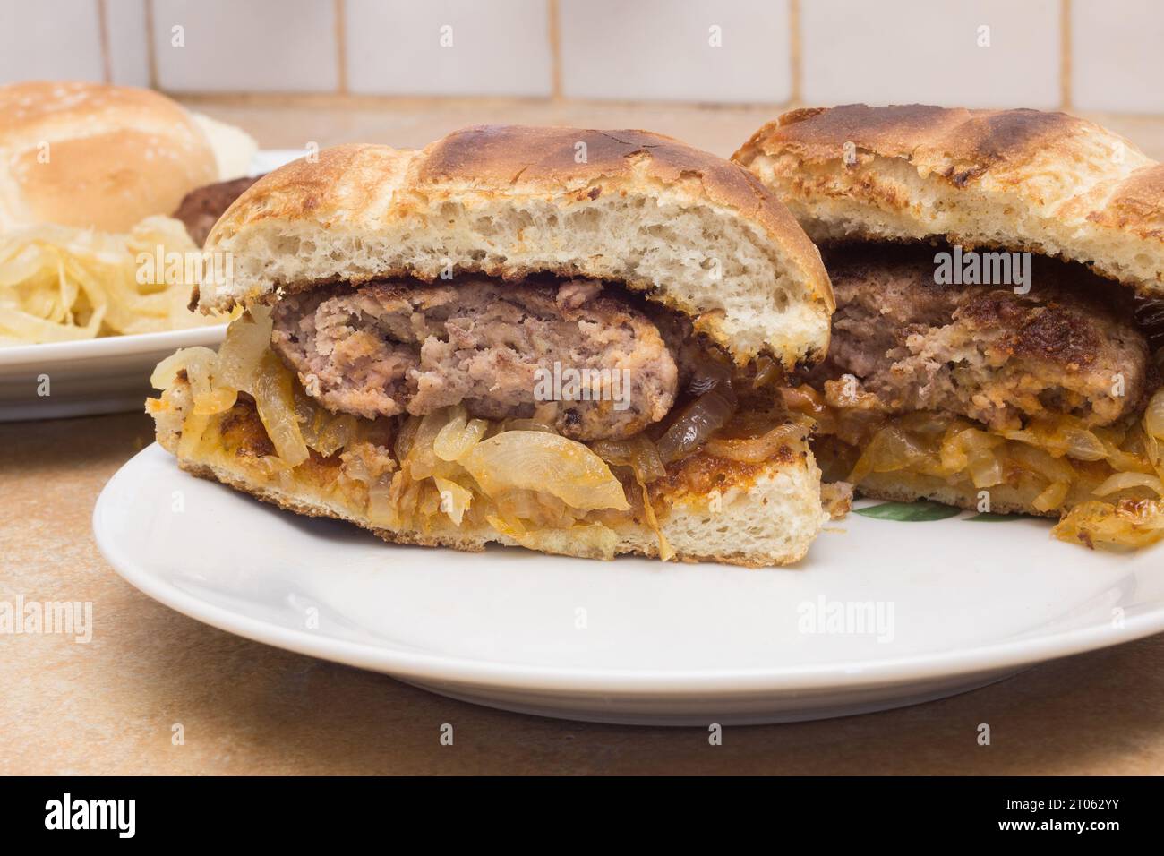 Un gros plan de deux moitiés d'un hamburger avec du pain grillé sur une assiette en céramique, placé sur un comptoir rustique, avec des oignons dorés en arrière-plan et un Banque D'Images