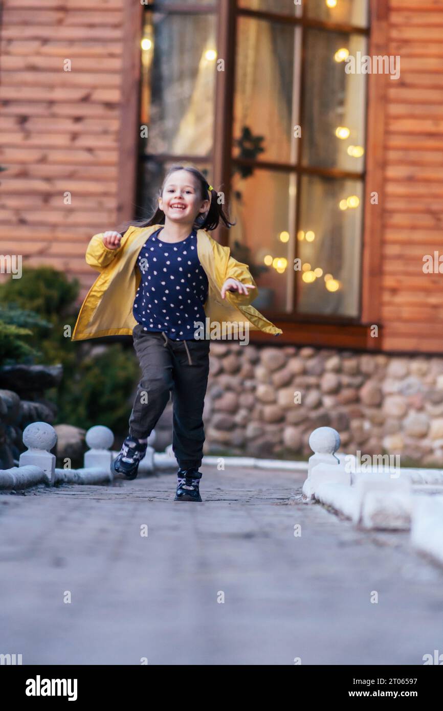 Glorieuse petite fille avec les cheveux bruns dans deux queues de cheval, dans la veste jaune courant de la maison et regardant la caméra avec le sourire heureux. Enfant qui va marcher Banque D'Images