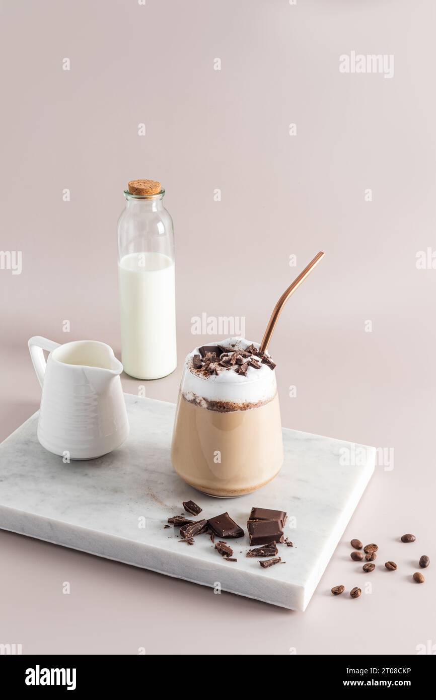 Un verre de café latte ou cappuccino avec des quartiers de chocolat se dresse sur un podium blanc en marbre avec une bouteille et un pichet de lait. Vue avant verticale Banque D'Images