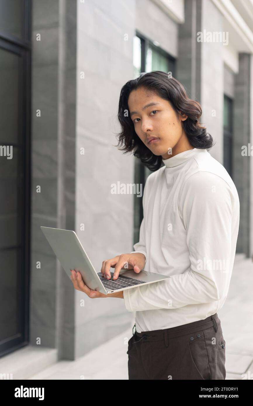 Jeune homme d'affaires asiatique utilisant un ordinateur portable à proximité de son espace de travail de bureau contre des bâtiments d'entreprise contemporains dans la ville. Banque D'Images