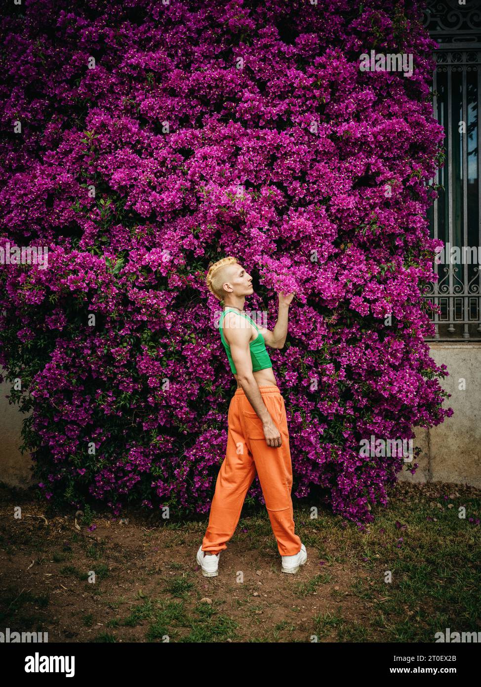 Personne non binaire avec un haut vert et un pantalon orange se tient devant un arbuste avec des fleurs Banque D'Images