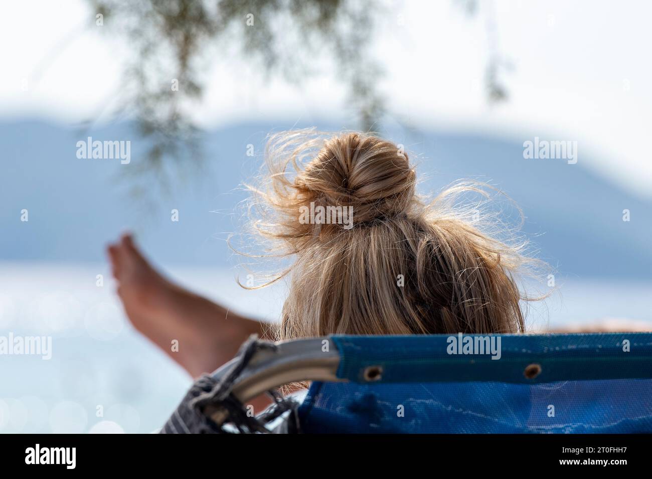 femme bronzer avec les cheveux dans un chignon ou topknot et un pied se dressant sur un transat dans le soleil méditerranéen en grèce Banque D'Images