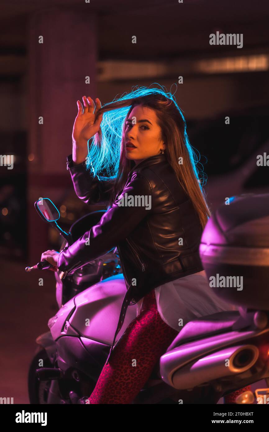 Photographie avec des néons bleus et roses sur une moto de derrière. Portrait d'un jeune modèle blond caucasien portant une veste en cuir noir Banque D'Images