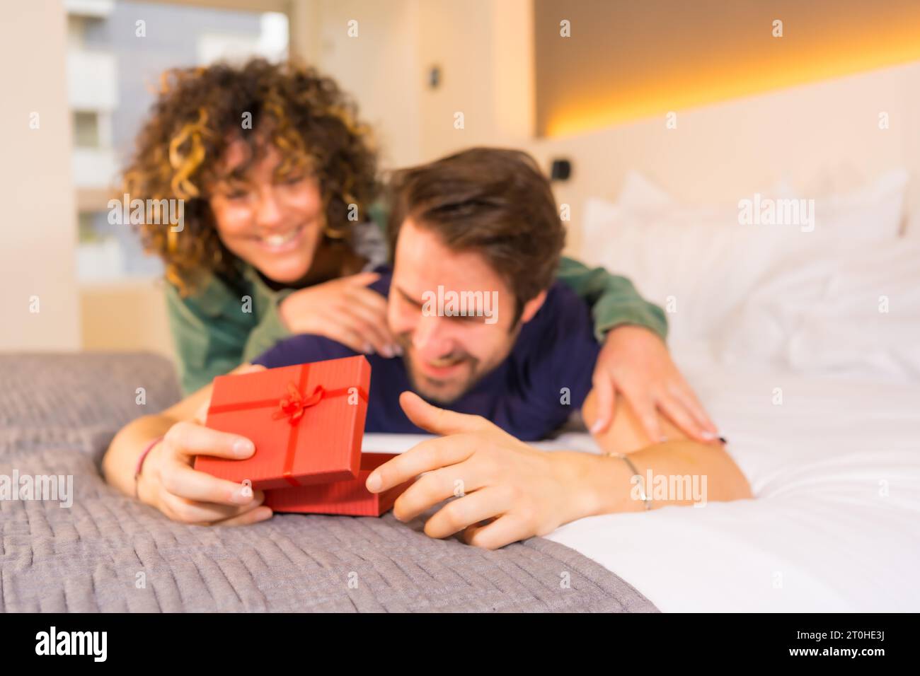 Saint Valentin, un jeune couple caucasien en pyjama sur le lit, la mariée donnant une belle surprise avec un cadeau au petit ami Banque D'Images Saint Valentin, un jeune couple caucasien en pyjama sur le lit, la mariée donnant une belle surprise avec un cadeau au petit ami Banque D'Images