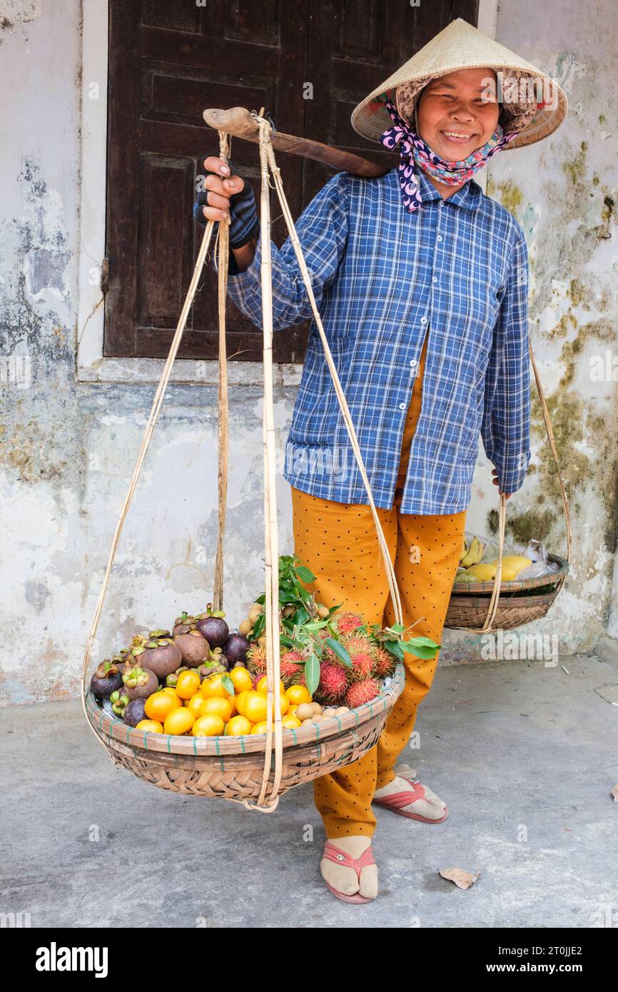 Hoi an, Vietnam. Femme portant des fruits tropicaux sur des poteaux d'épaule. Banque D'Images