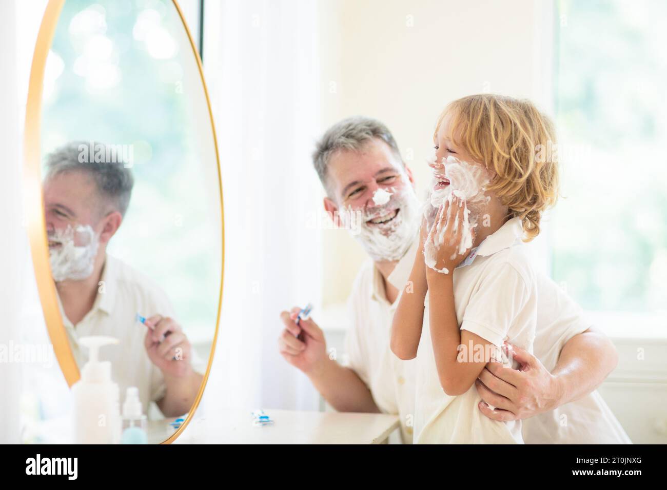 Père et enfant dans la salle de bain regardant dans le miroir. Papa se rasant dans la salle de bain ensoleillée blanche, petit garçon jouant avec la mousse. Jeune homme enseignant à son fils à se raser. Banque D'Images