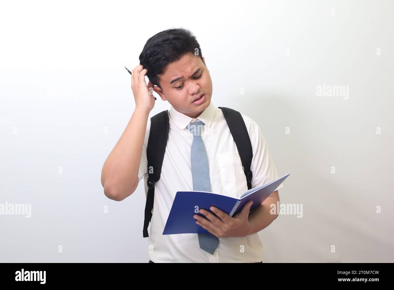 Lycéen indonésien portant un uniforme de chemise blanche avec cravate grise écrivant sur le carnet de notes à l'aide d'un stylo avec une expression agacée et frustrée. Banque D'Images