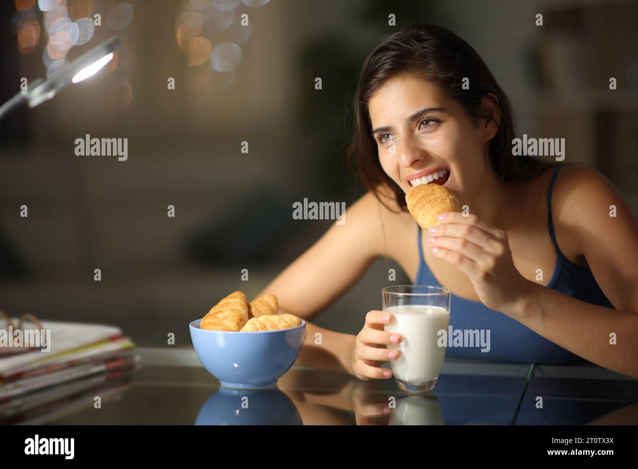 Femme heureuse mangeant des croissants et du lait assis dans la nuit à la maison Banque D'Images