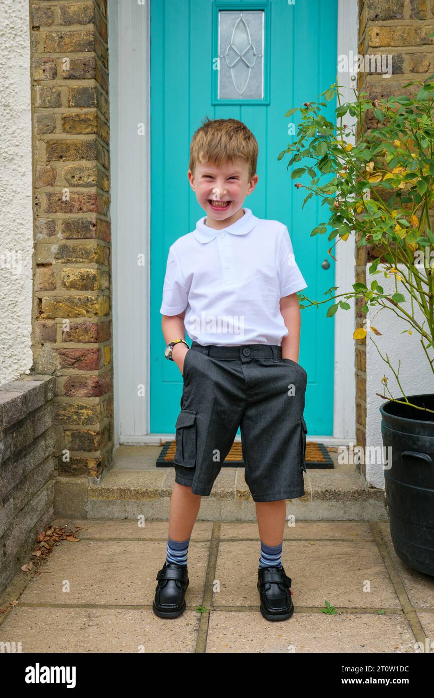 Heureux et excité petit garçon de 4 ans devant la porte d'entrée de sa maison sur son premier jour d'école portant son uniforme et un pantalon court, Royaume-Uni Banque D'Images